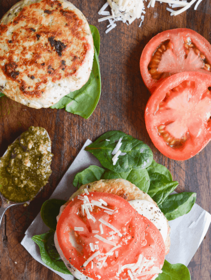 two chicken pesto burgers on a brown cutting board with sliced tomatoes in one corner and a spoon of pesto in another