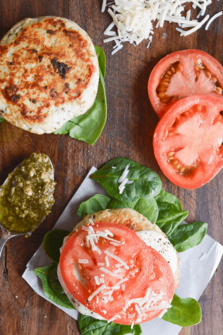 two chicken pesto burgers on a brown cutting board with sliced tomatoes in one corner and a spoon of pesto in another