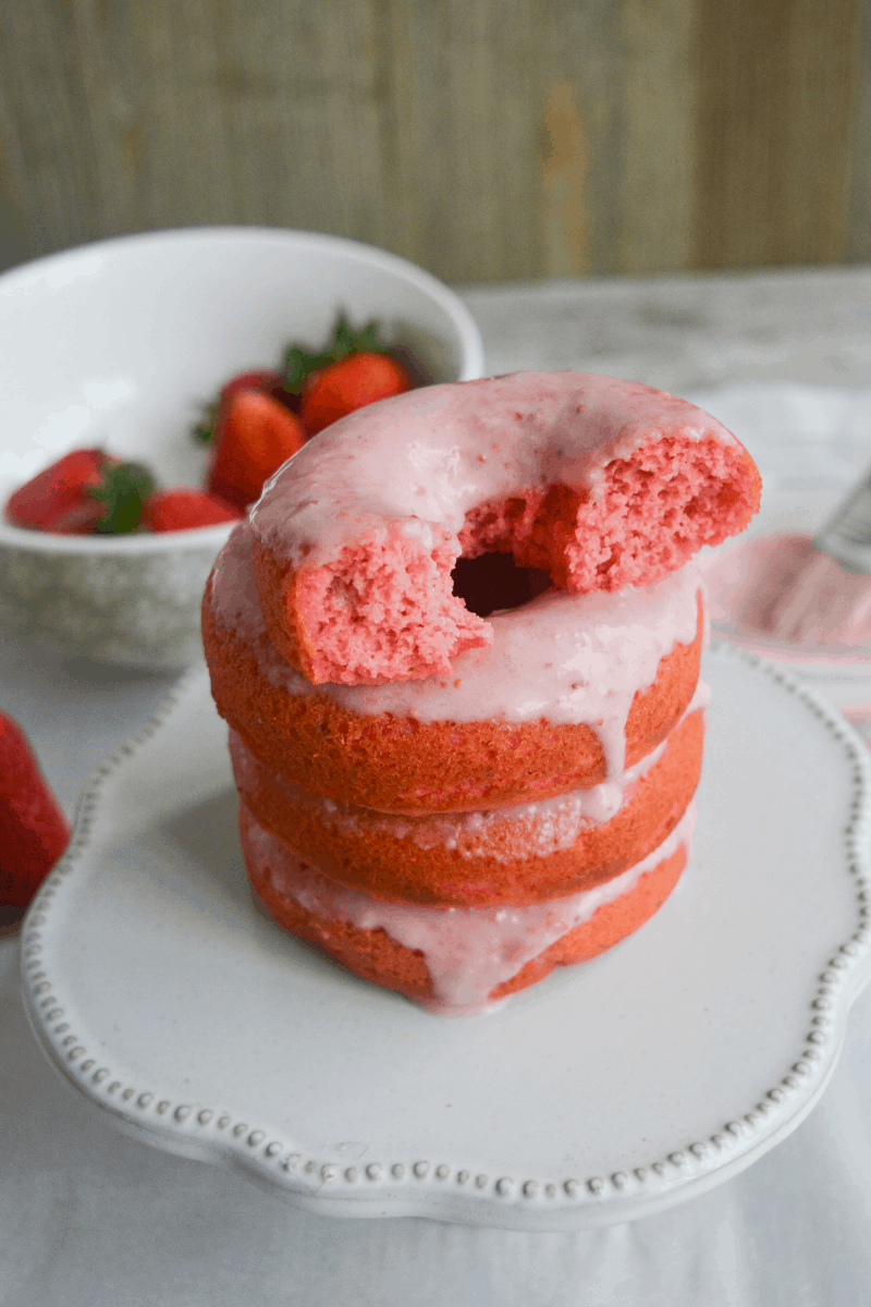 four glazed keto strawberry donuts stacked on white stand with bowl of strawberries in background