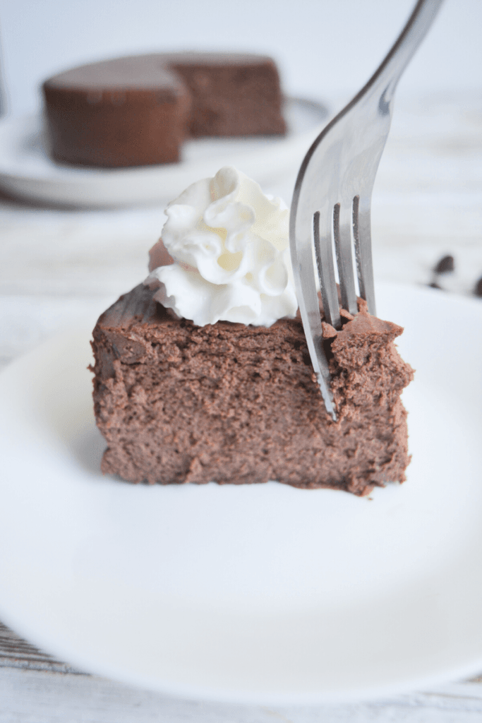 slice of chocolate cheesecake on a white plate with a dollop of whipped cream on top. a fork is taking a small portion out of the slice.