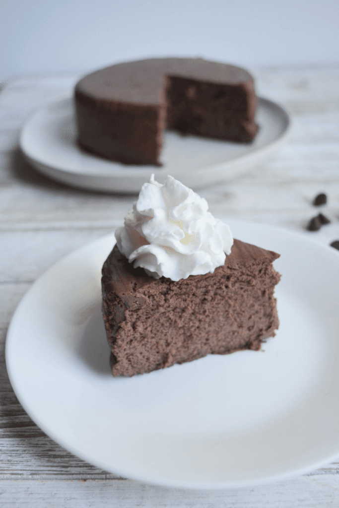 slice of instant pot keto cheesecake in the foreground on a white plate with a dollop of whipped cream on top. the remainder of the cheesecake sits in the backgroudn on a different white plate.