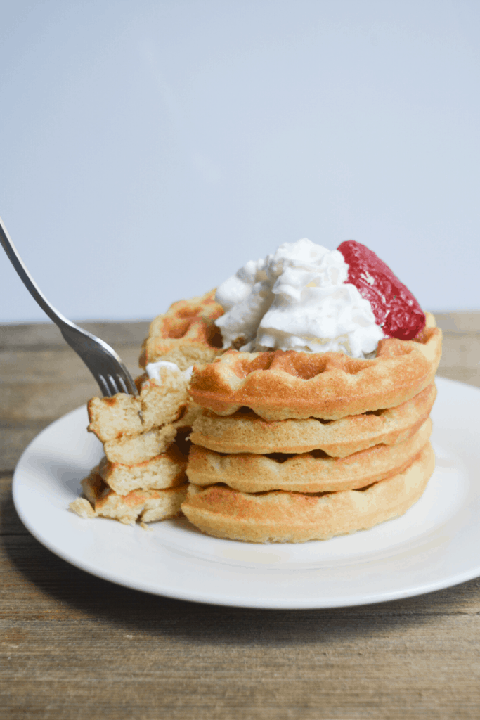 stack of keto waffles on a white plate topped with whipped cream and a strawberry. a fork is taking a slice of the waffle stack
