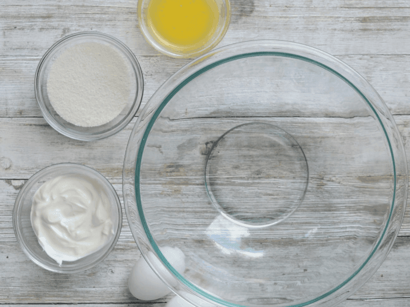 empty mixing bowl sitting beside bowls of sour cream, melted butter, vanilla extract, and granular swerve, respectively and eggs