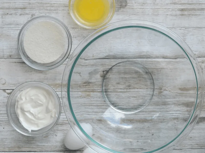 empty mixing bowl sitting beside bowls of sour cream, melted butter, vanilla extract, and granular swerve, respectively and eggs