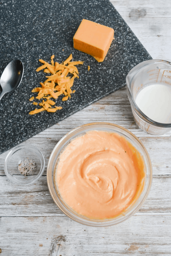 cheese sauce sitting in a bowl surrounded by ingredients, heavy whipping cream, shredded cheddar cheese and salt and pepper