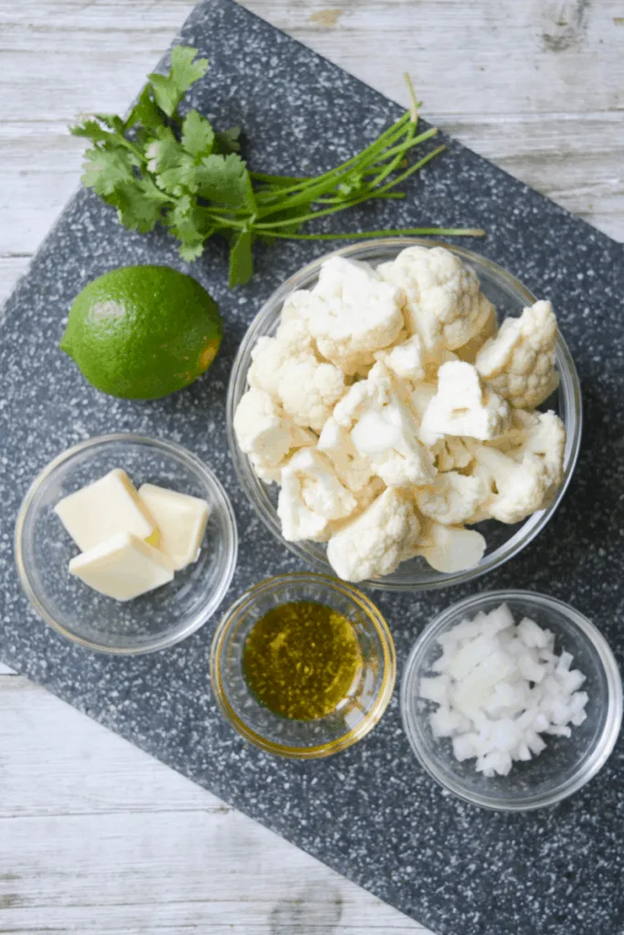 ingredients for cauliflower rice sitting on a grey speckled cutting board. these ingredients include: cilantro, lime, butter, olive oil, onion, and cauliflower florets.