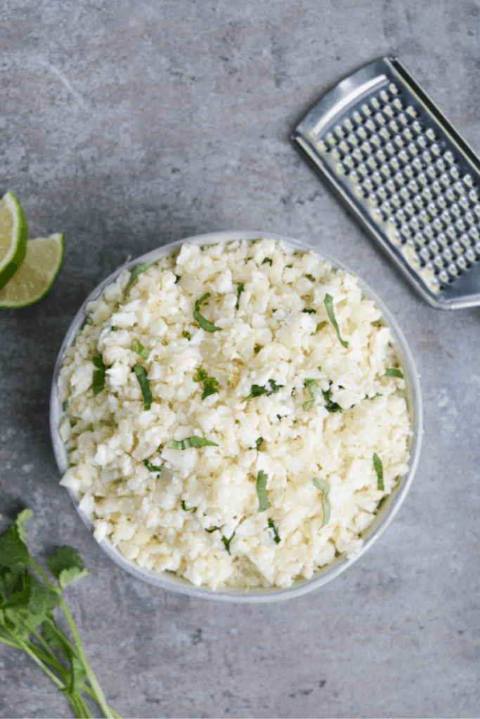 bowl of cauliflower race sitting surrounded by fresh lime slices, fresh cilantro, and a zester