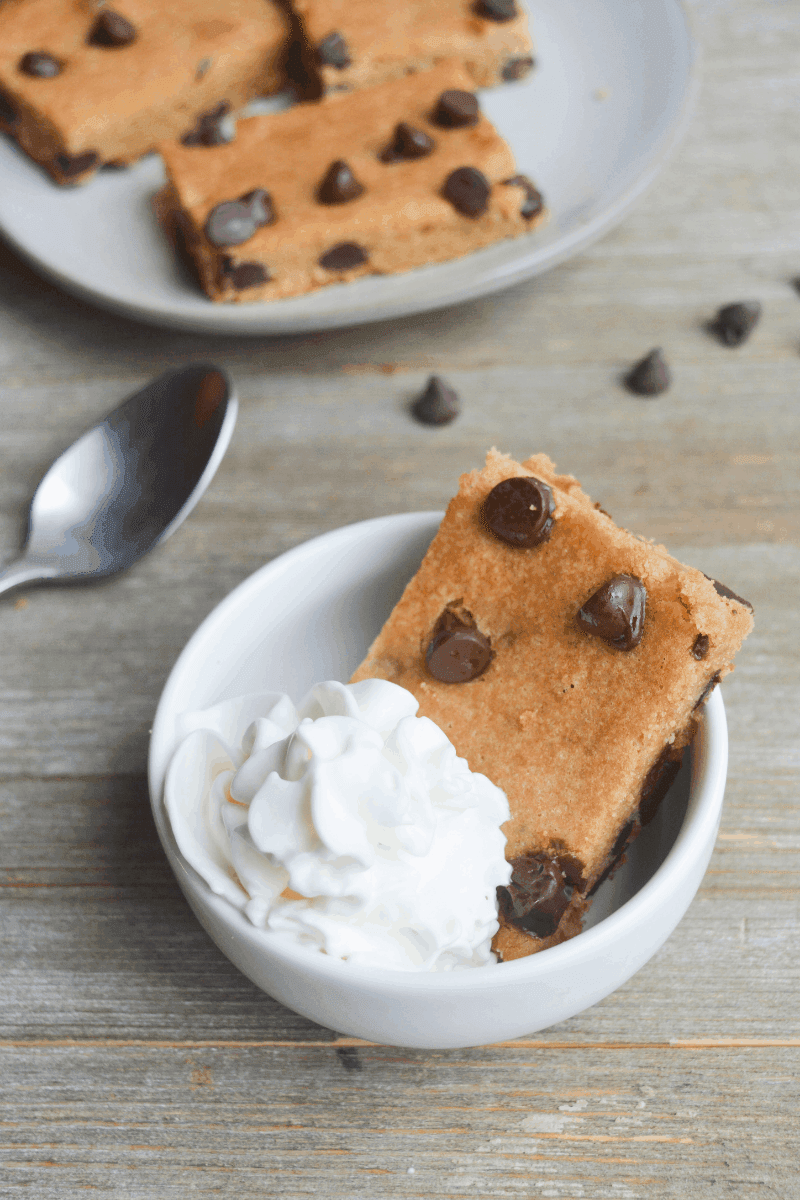 keto chocolate chip cookie bar in a small bowl with whipped cream. in the background there is a plate of cookie bars