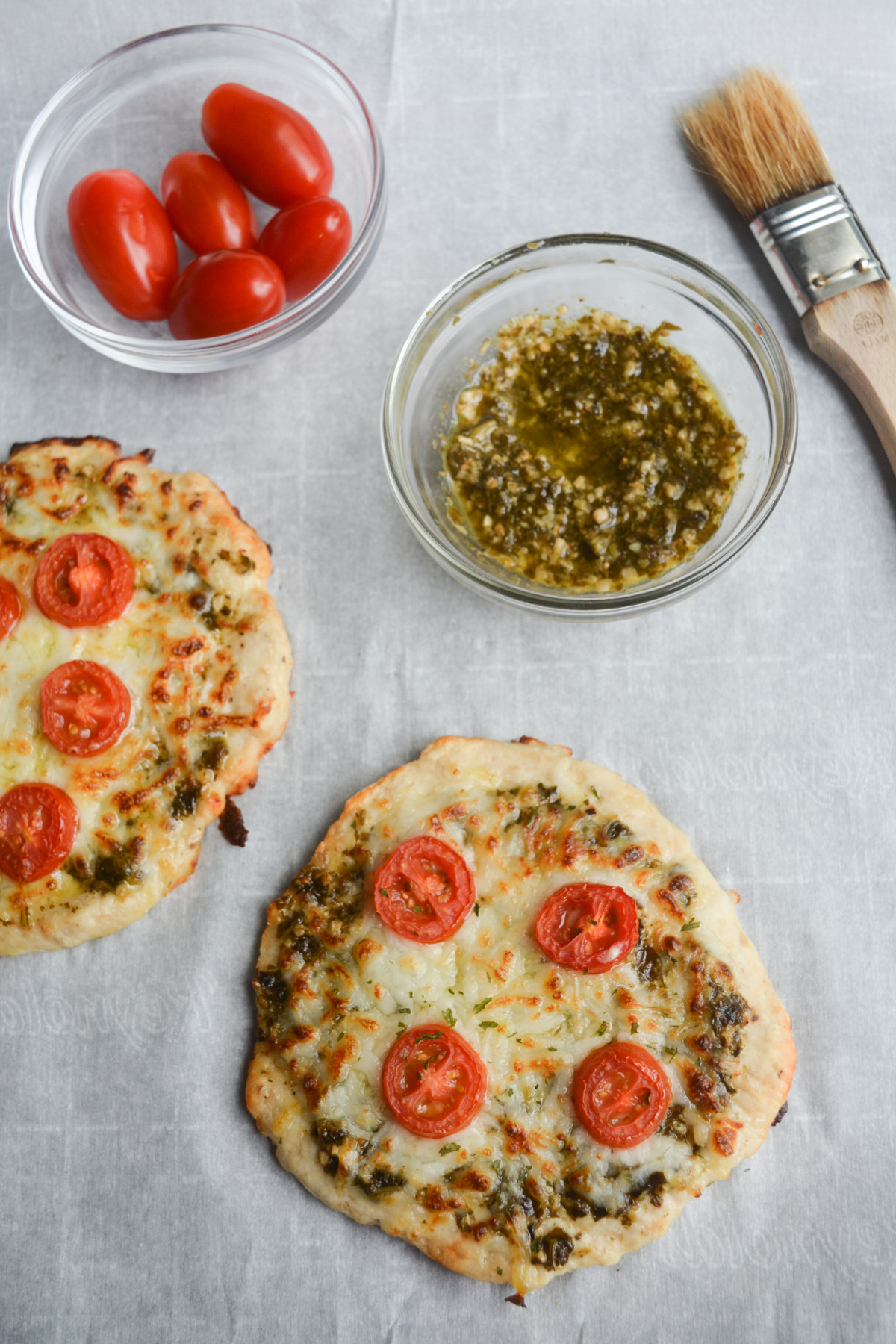 Keto Chicken Pesto Pizzas, topped with sliced grape tomatoes, sitting on a piece of parchment paper, with a small bowl of basil pesto in the upper right hand corner.