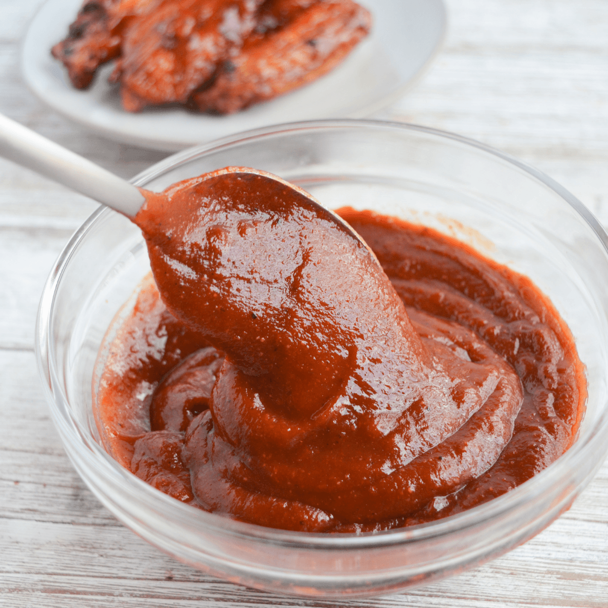 bowl of keto bbq sauce with a spoon scooping some out. sauced chicken wings sit in the background.