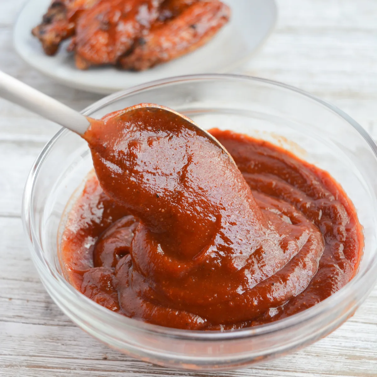 bowl of keto bbq sauce with a spoon scooping some out. sauced chicken wings sit in the background.