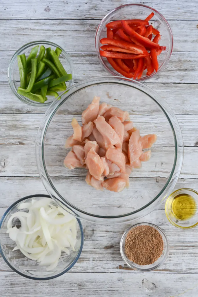 chicken fajitas ingredients, including sliced chicken breast, sliced red bell pepper, sliced green bell pepper, sliced yellow onion, fajita seasoning, and olive oil.