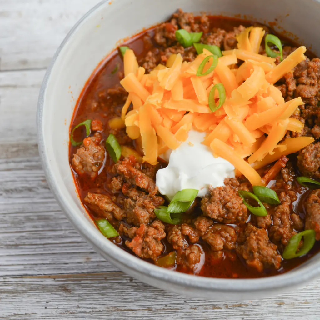 close up on a bowl of keto chili topped with green onions, sour cream, and shredded cheddar cheese