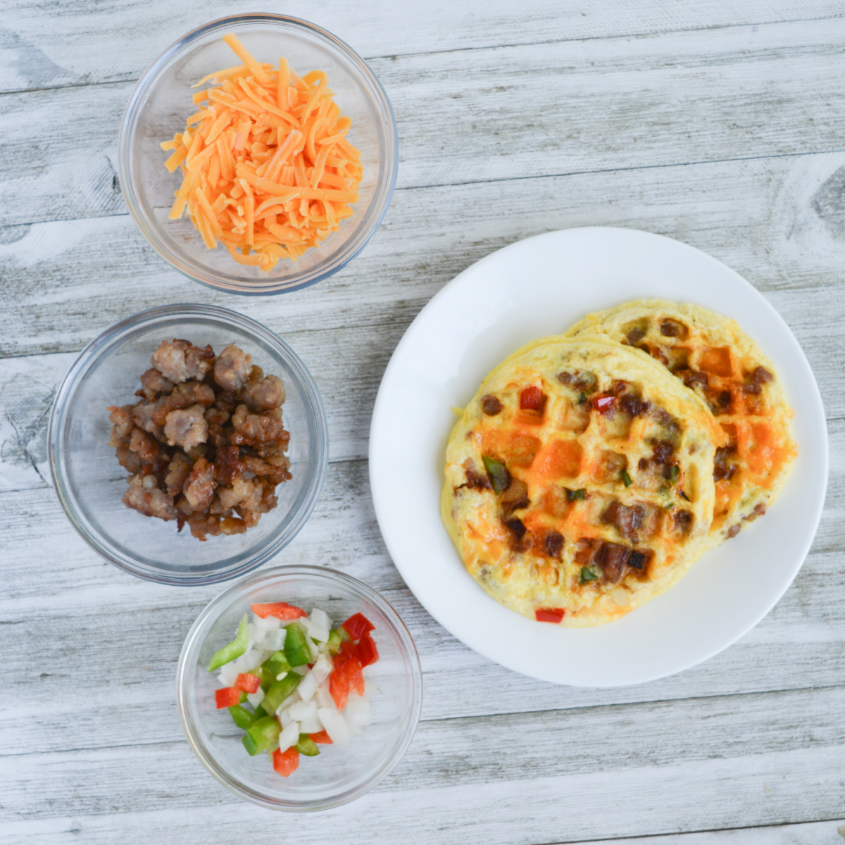 overhead view of two cooked waffle omelettes on a plate and three bowls filled with shredded cheddar, crumbled breakfast sausage, and diced vegetables