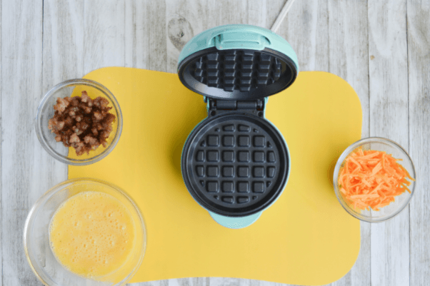 three bowls, containing whisked eggs, crumbled breakfast sausage and shredded cheddar cheese, respectively. there is also an open mini waffle iron