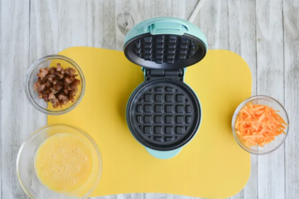 three bowls, containing whisked eggs, crumbled breakfast sausage and shredded cheddar cheese, respectively. there is also an open mini waffle iron