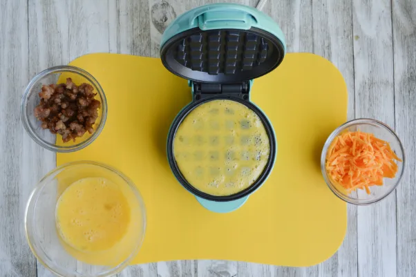 three bowls, containing whisked eggs, crumbled breakfast sausage and shredded cheddar cheese, respectively. there is also an open mini waffle iron and it is ⅔ of the way full with whisked egg