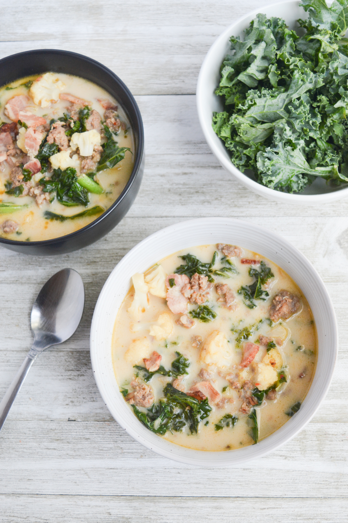 one black bowl of low carb zuppa toscana soup, one white bowl of the same, and a white bowl of fresh kale.