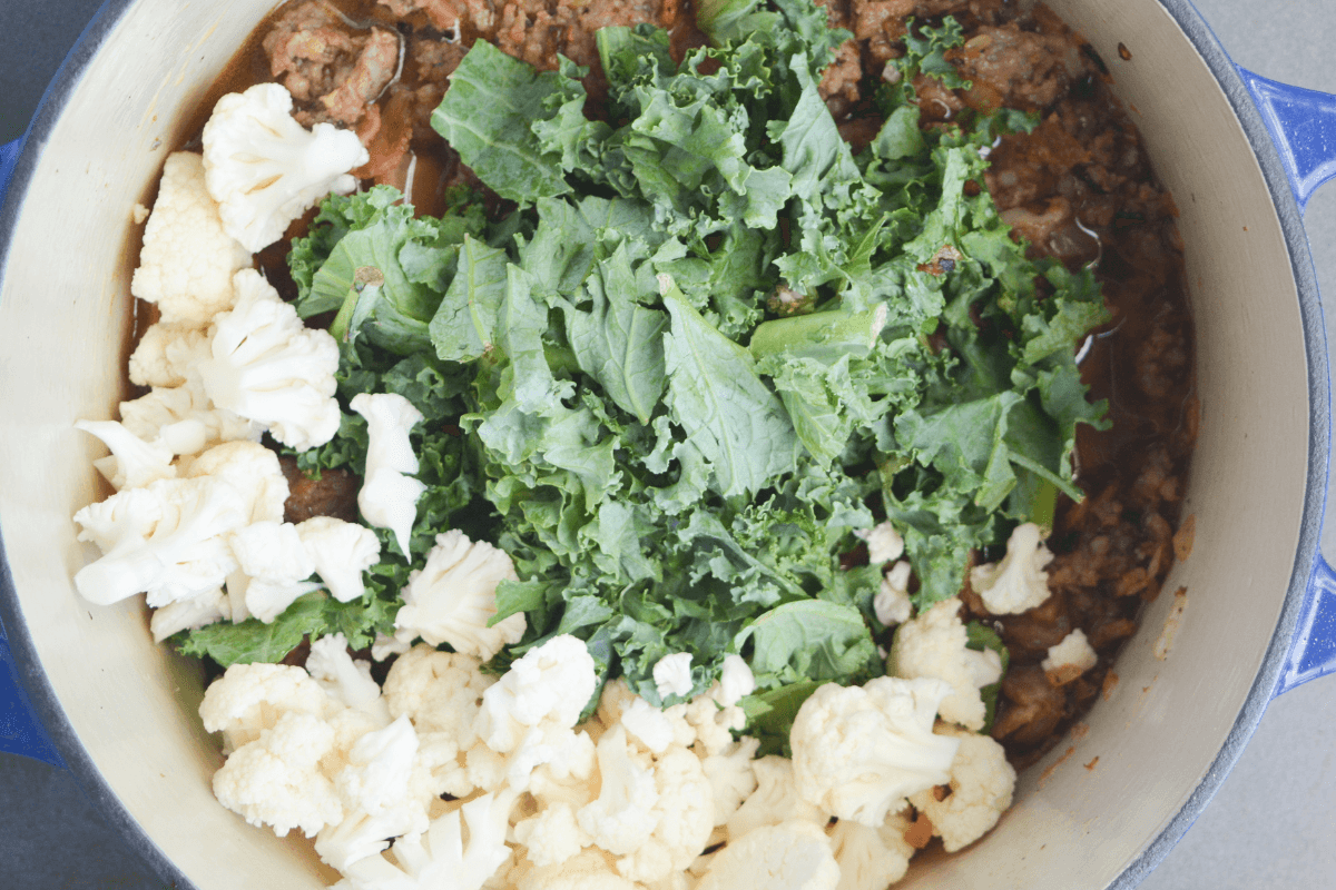 chopped cauliflower and chopped kale on top of Italian sausage in a large pot.