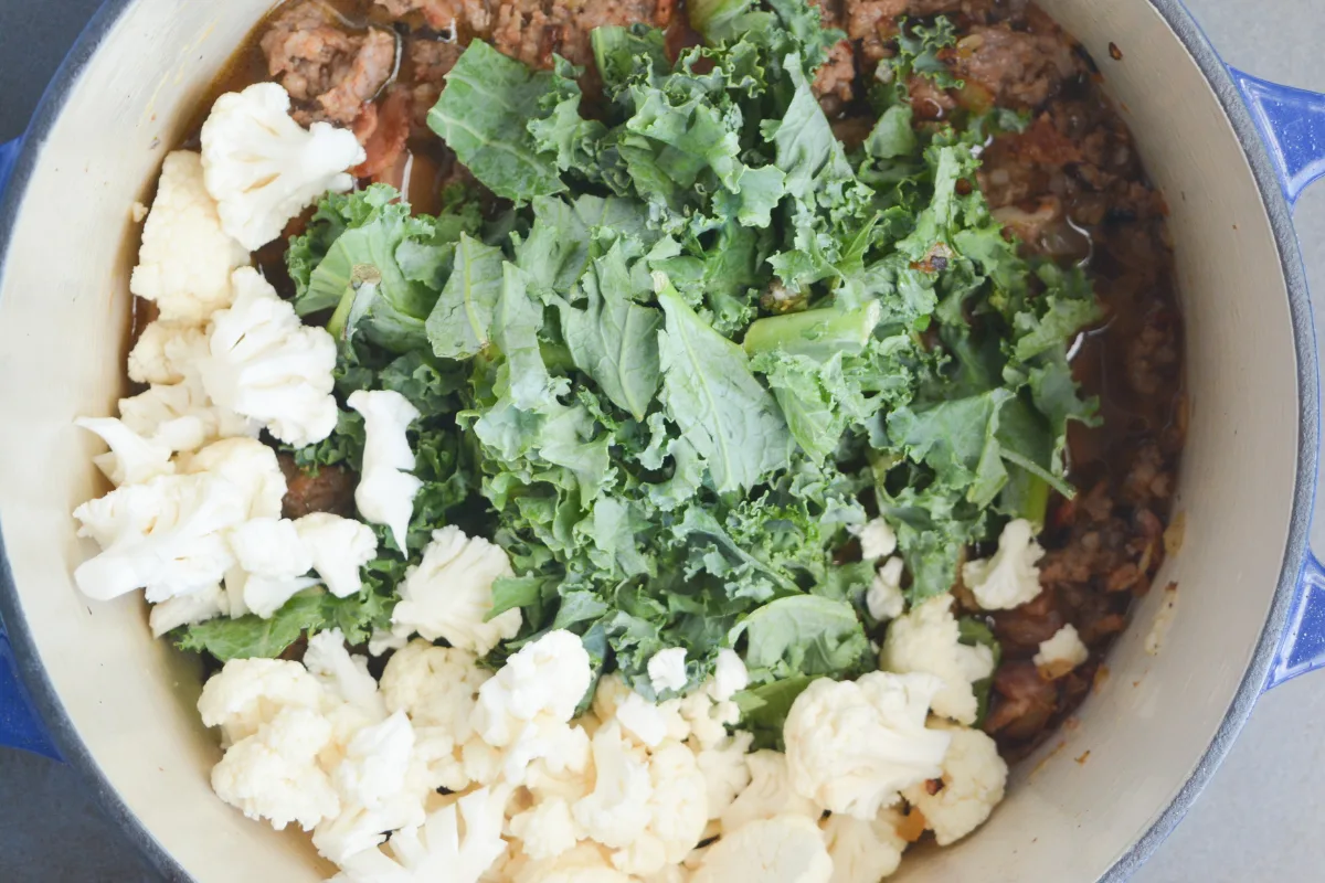 chopped cauliflower and chopped kale on top of Italian sausage in a large pot.