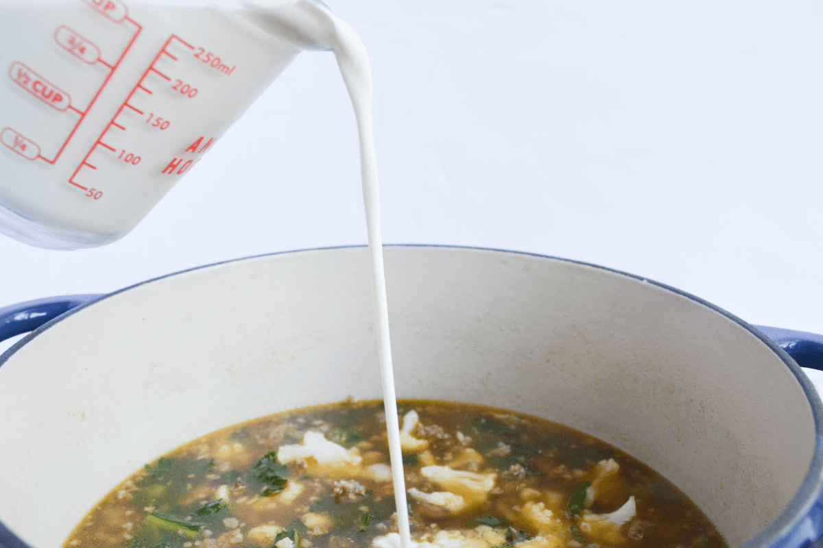 heavy cream being added to a large pot of low carb zuppa toscana ingredients.