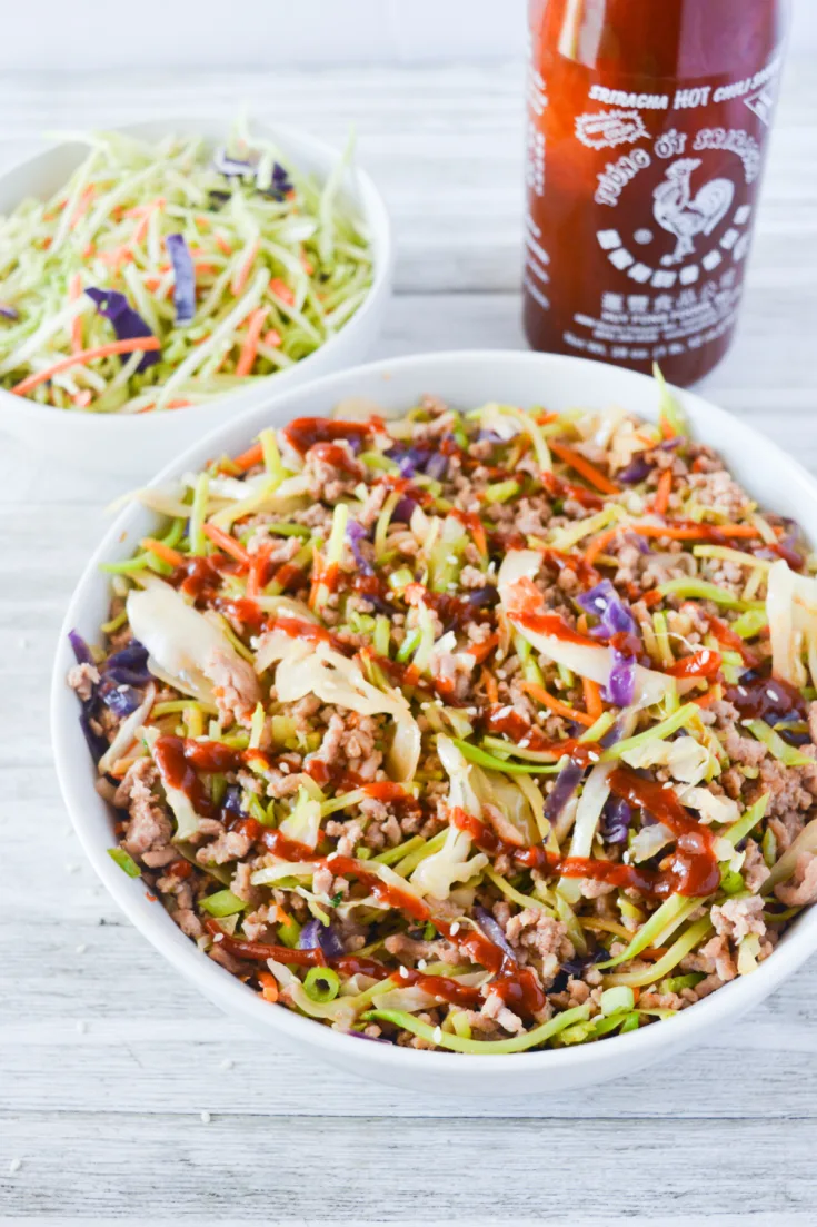 White bowl on wooden tabletop with green vegetables, carrots, pork and a drizzle of sriracha